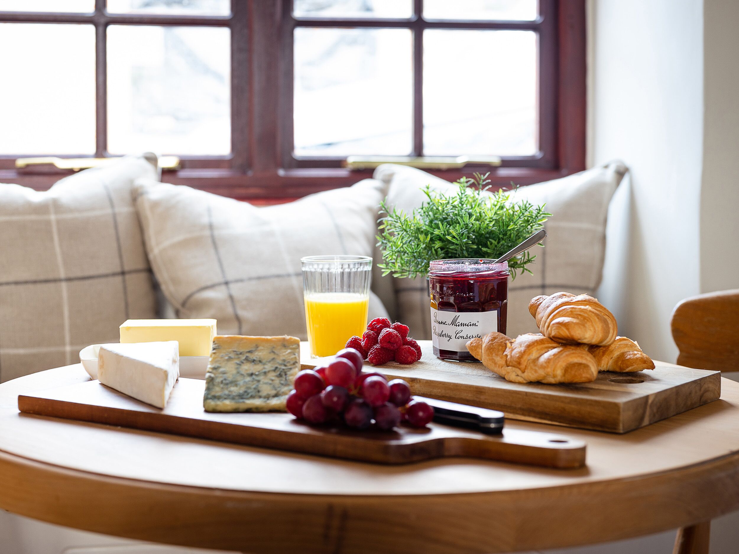 Una escena de desayuno con queso, uvas, un vaso de zumo de naranja, un frasco de mermelada y croissants sobre una tabla de madera.