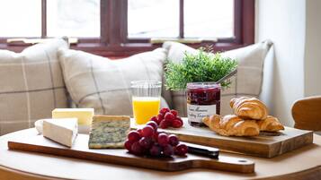 Una escena de desayuno con queso, uvas, un vaso de zumo de naranja, un frasco de mermelada y croissants sobre una tabla de madera.