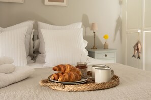 A neatly made bed with white bedding, a basket with croissants and a jar of jam, and a bedside table with a lamp and a vase of flowers.