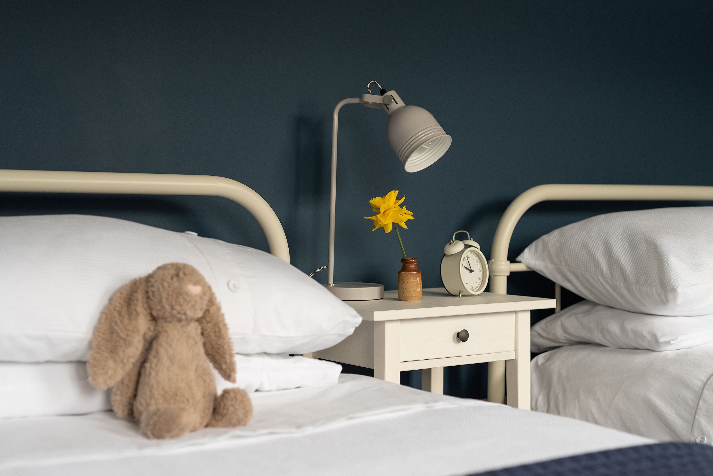 A neatly made bed with white bedding, a teddy bear, a bedside lamp, a flower vase, and a clock on a bedside table.
