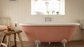 A bathroom with a freestanding bathtub, a wooden stool, a candle, and a window with a curtain.