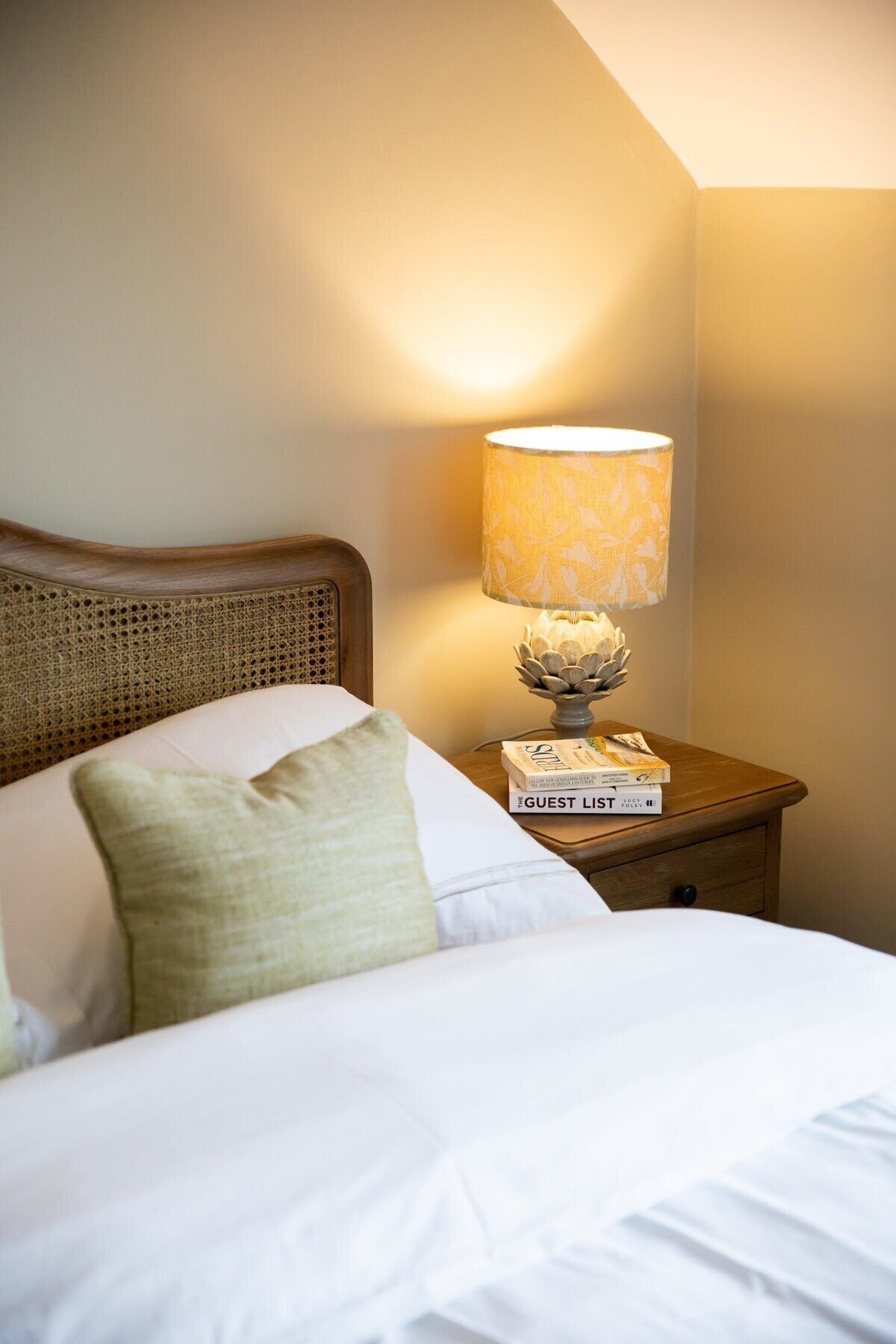 Una cama bien hecha con sábanas blancas y una única almohada verde. Un mesita de noche de madera con una lámpara y una pila de libros está al lado de la cama.