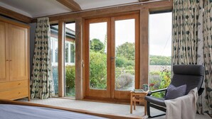 A bedroom with a bed, a chair, a small table, and a view of a garden through a sliding door. - Charming Cottage for 10 in Heart of South Downs (Eartham)