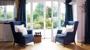 A room with two blue armchairs, a wicker side table, and a view of greenery through a glass door. - Charming Cottage for 10 in Heart of South Downs (Eartham)