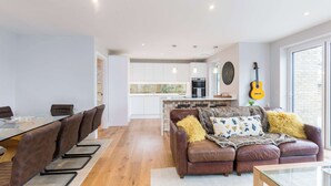 A modern living room with a dining area, a kitchen in the background, and a large window. - Stylish new home with parking - king beds, garden (Chichester)