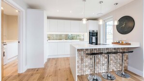 A modern kitchen with white cabinetry, a central island with stools, pendant lighting, and a large round clock on the wall. - Stylish new home with parking - king beds, garden (Chichester)