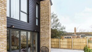 A modern house with a brick facade, large sliding glass doors, and a patio area with a wooden fence and a chair. - Stylish new home with parking - king beds, garden (Chichester)