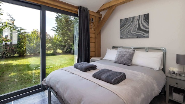 A bedroom with a large window, a bed with pillows, a nightstand, and a painting on the wall. - Rural Lodge Retreat with Hot Tub (Chichester)