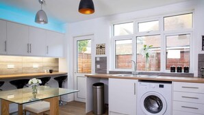A modern kitchen with white cabinets, a glass table, a washing machine, and a trash bin. - Spacious House for 10 Near Town & Beach (Bognor Regis)
