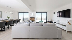 Een moderne woonkamer met een grote bank, een eethoek met stoelen, een aan de muur bevestigde televisie en grote ramen.