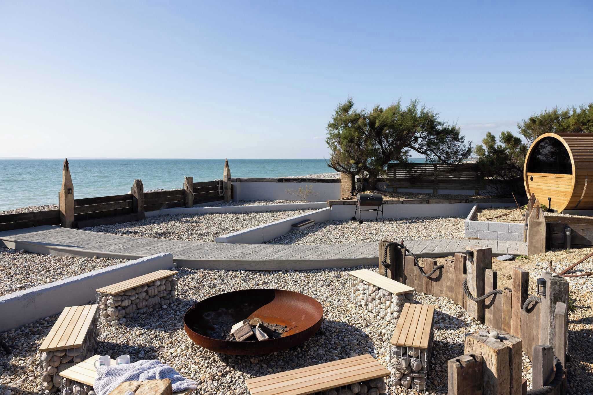 A beachside relaxation area with a fire pit, wooden benches, and a wooden barrel structure.