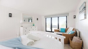 Een slaapkamer met een groot bed, een kaptafel, een rieten stoel en uitzicht op de natuur door een schuifdeur.