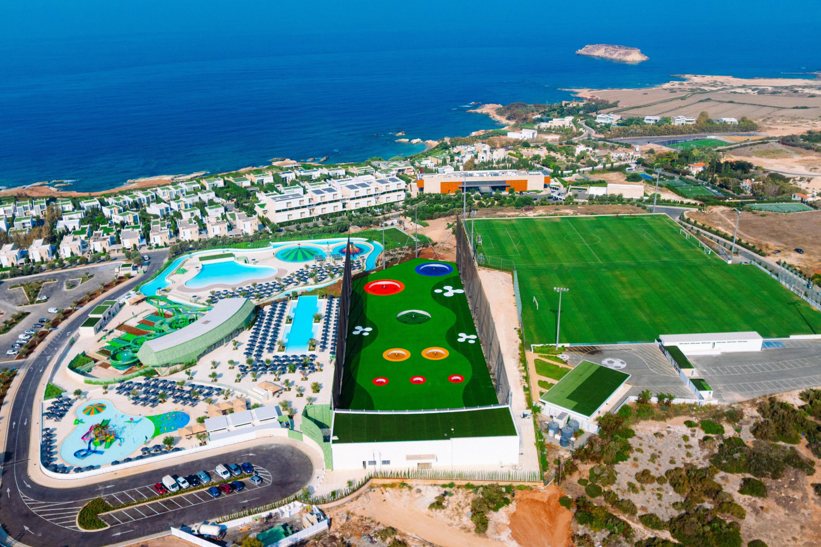 An aerial view of a resort with a swimming pool, sports facilities, and a beach area.