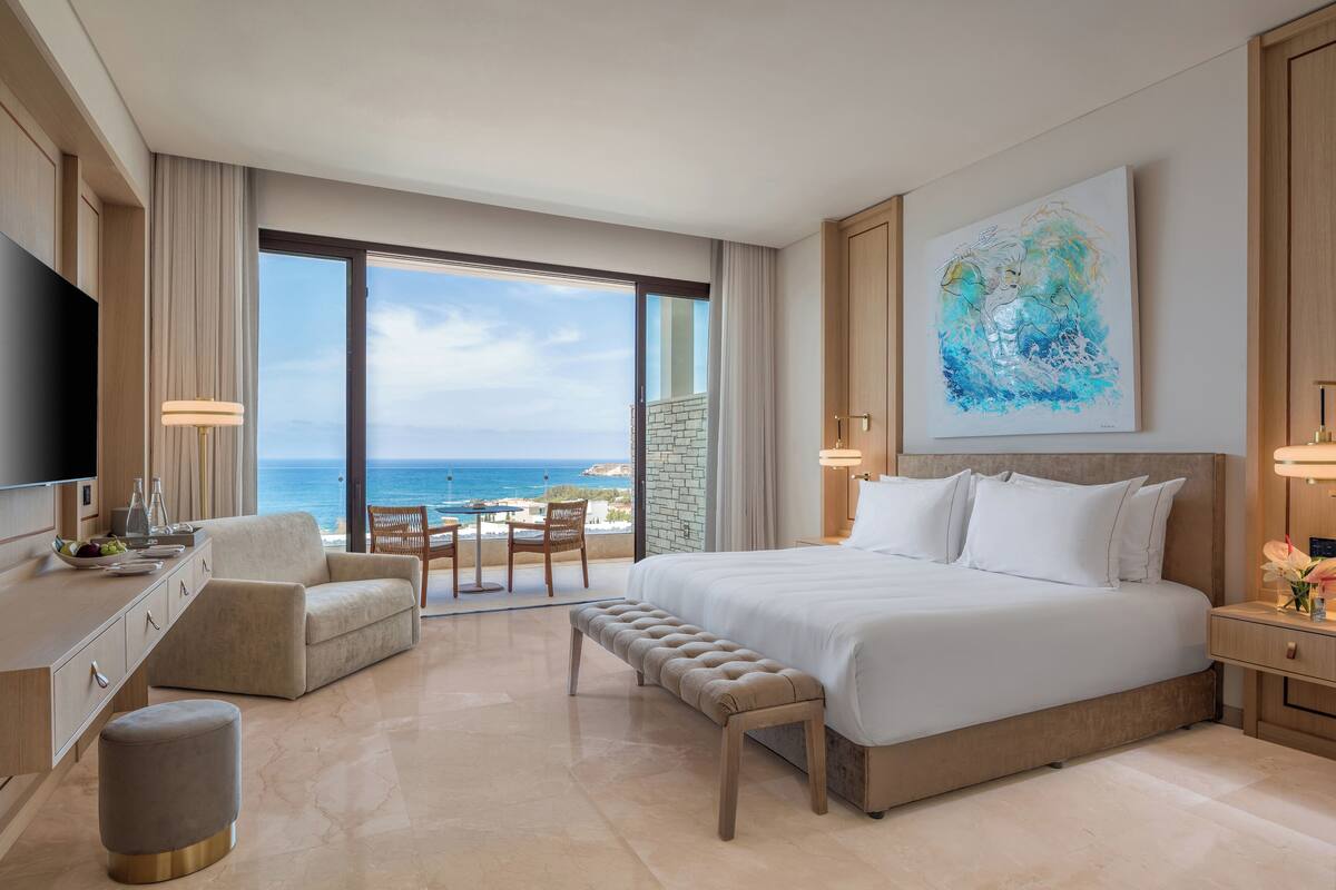 Une chambre d’hôtel moderne dotée d’un grand lit, d’un coin salon avec vue sur l’océan, d’une télévision à écran plat et d’un coin repas avec une table et des chaises.