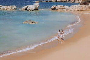 Une plage à l’eau claire, avec des formations rocheuses, où un couple se promène le long du rivage.