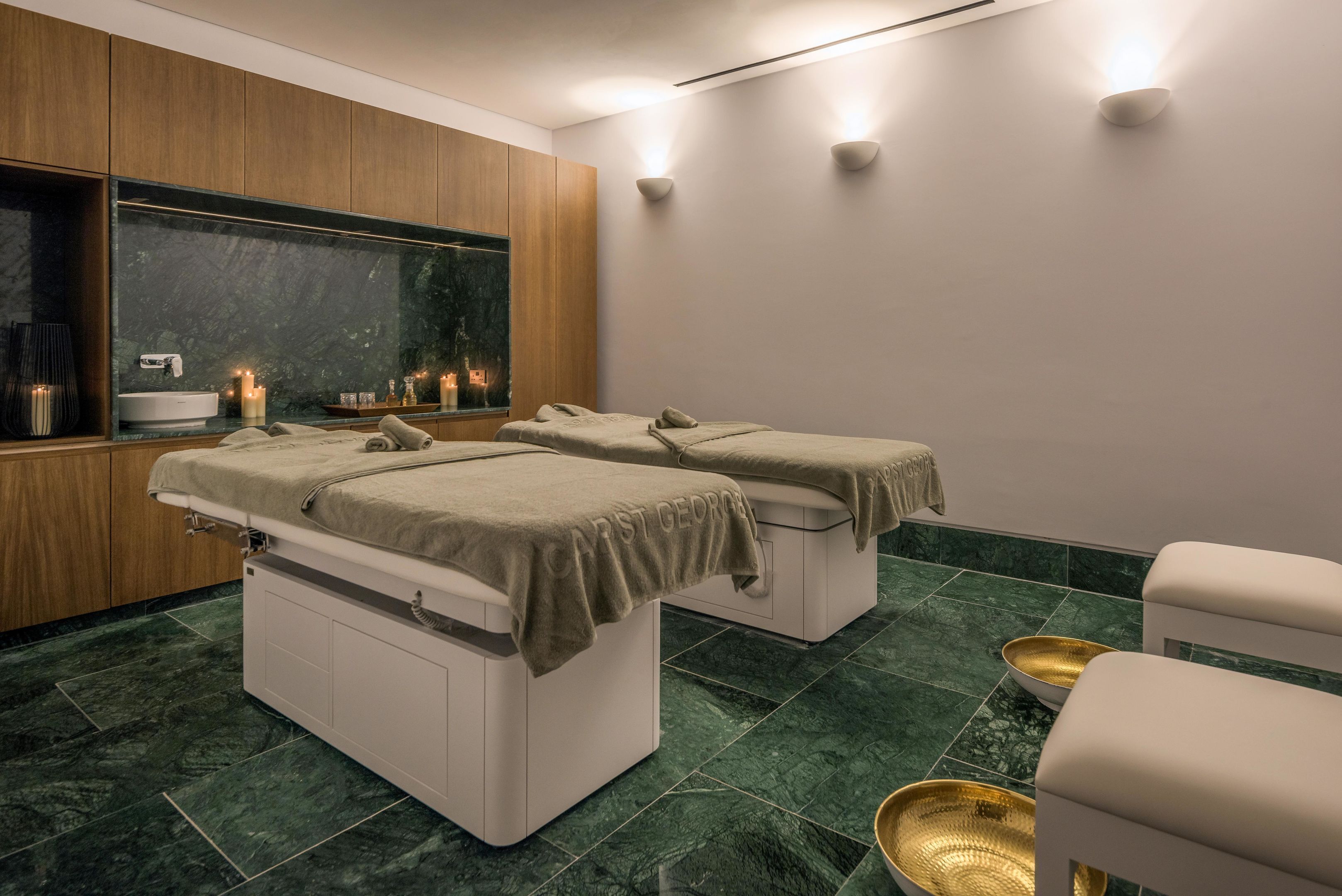 A spa room with two treatment beds, a large mirror, a sink, candles, and a wooden cabinet.