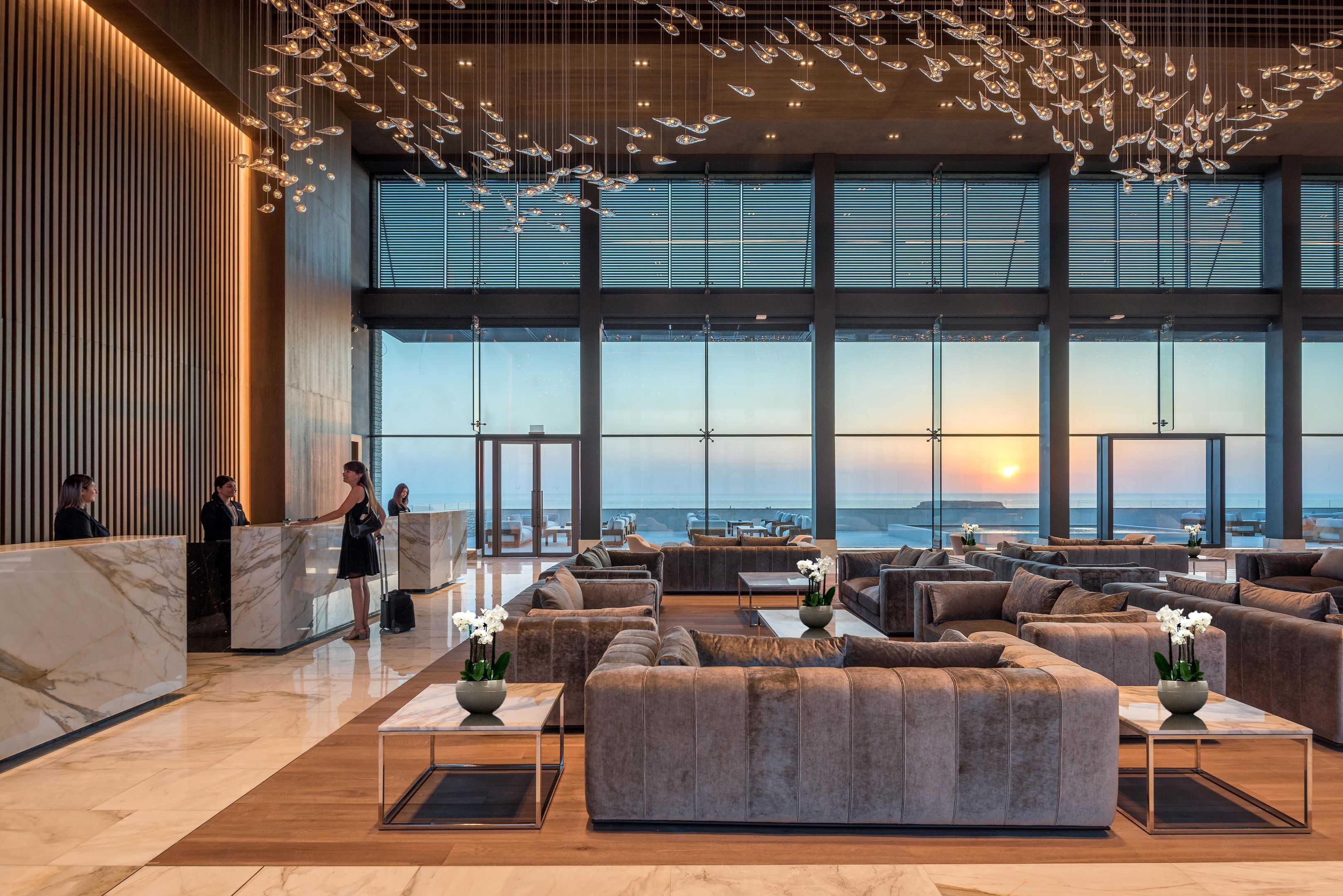 A spacious lobby with a reception desk, seating area, and large windows offering a view of the sunset.