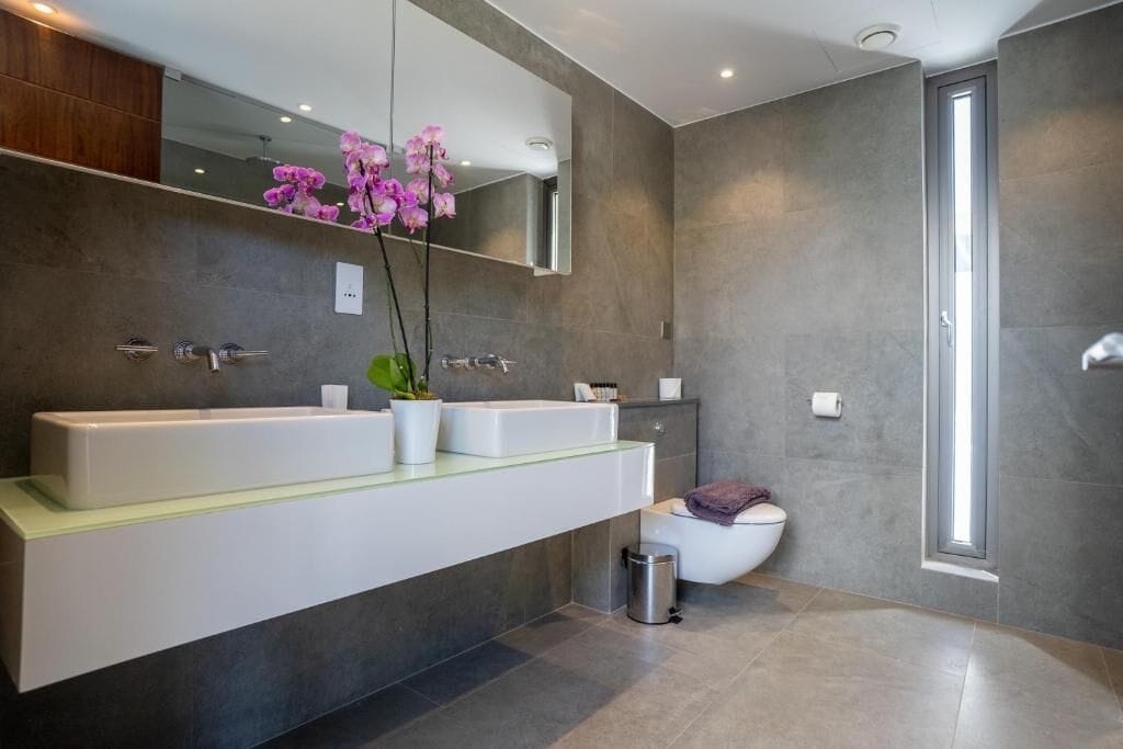 A modern bathroom with a large mirror, two sinks, a toilet, and a trash bin.