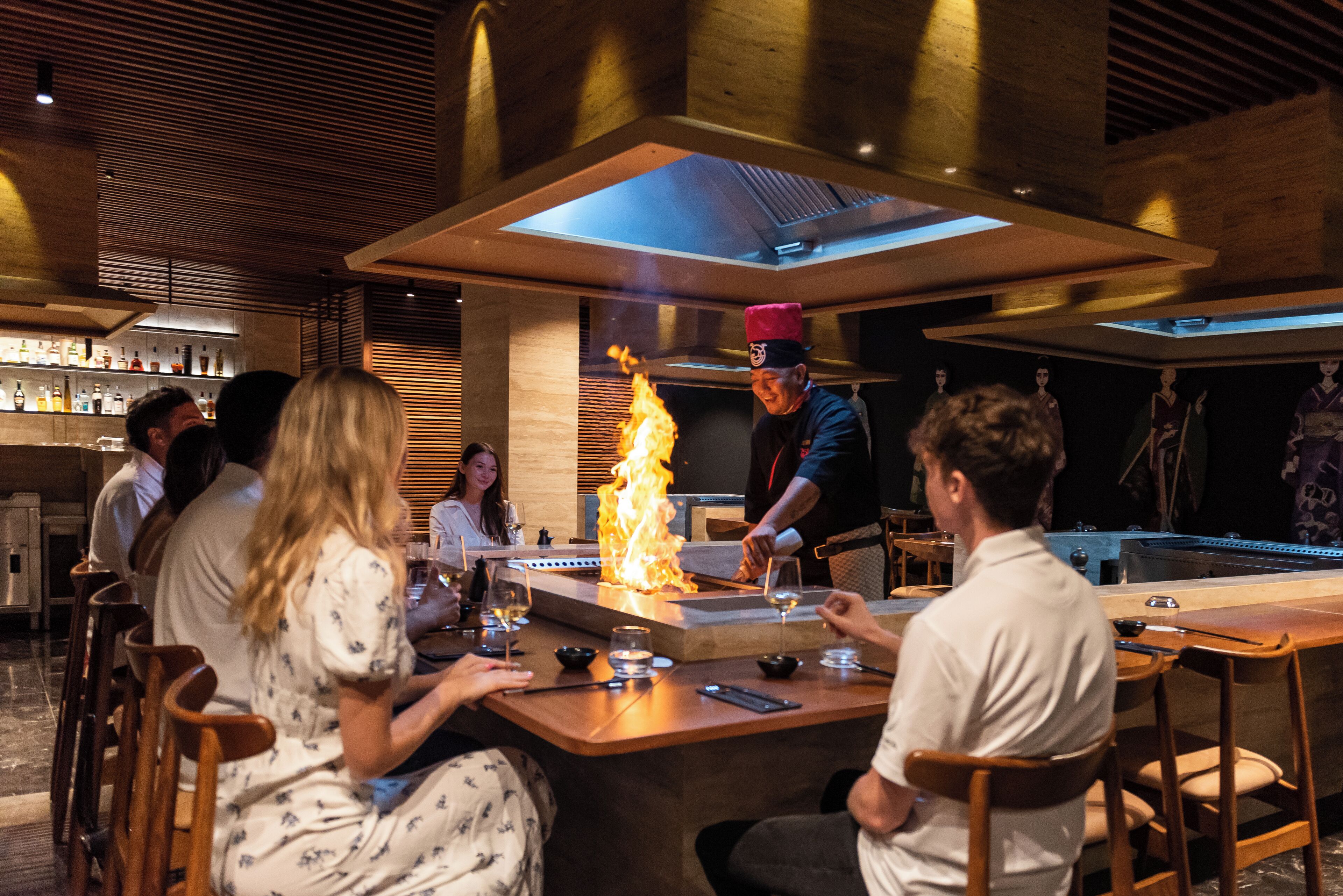 A restaurant with a central fire feature, wooden furniture, and a modern interior design.