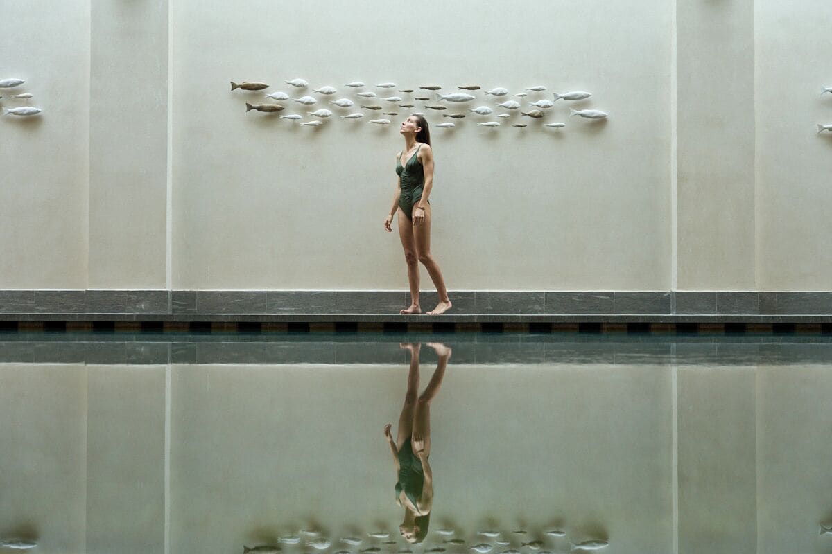 A person standing by a reflective pool with a wall of fish sculptures.
