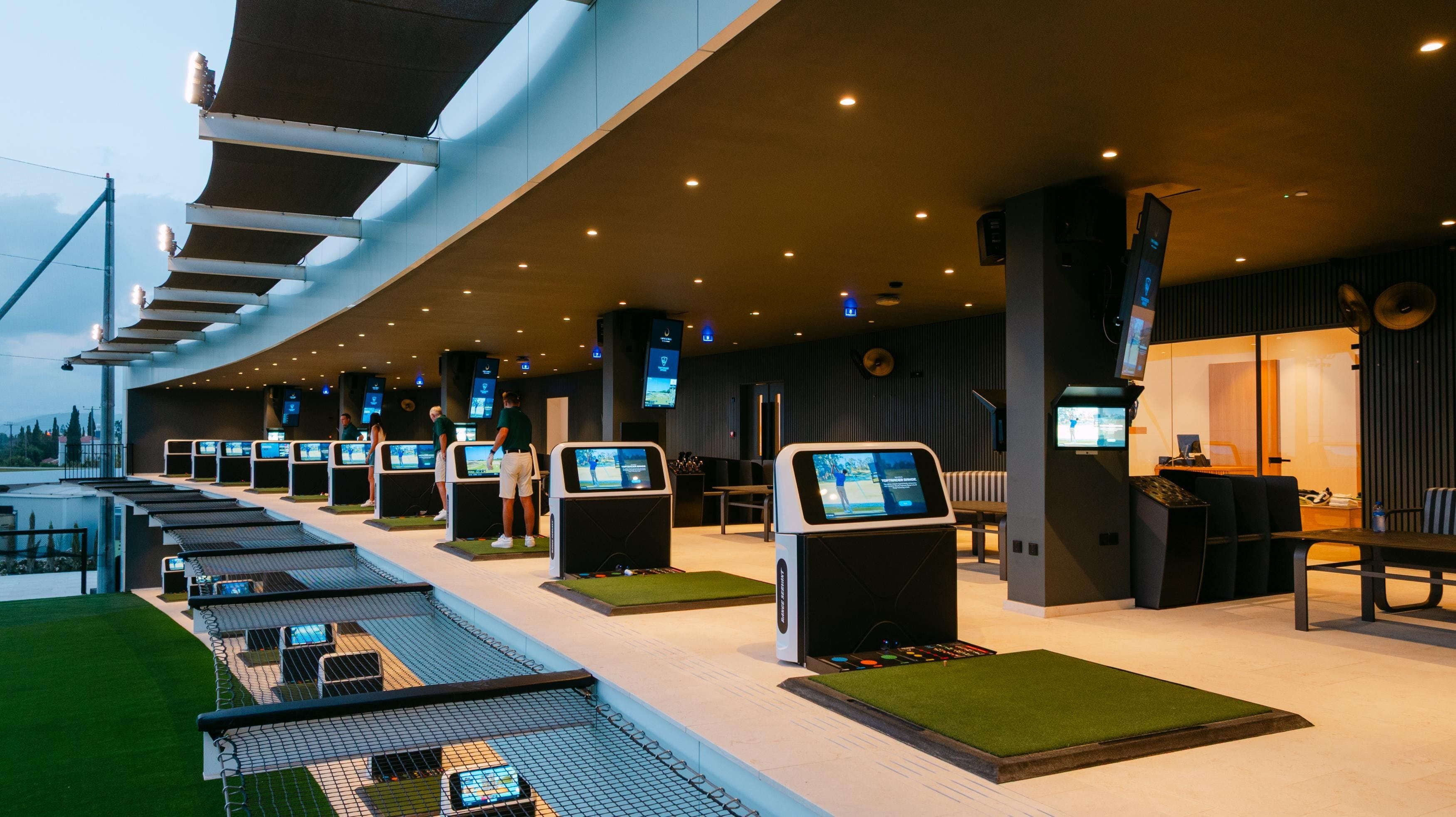 A modern indoor golf driving range with multiple stations, a seating area, and a pool.