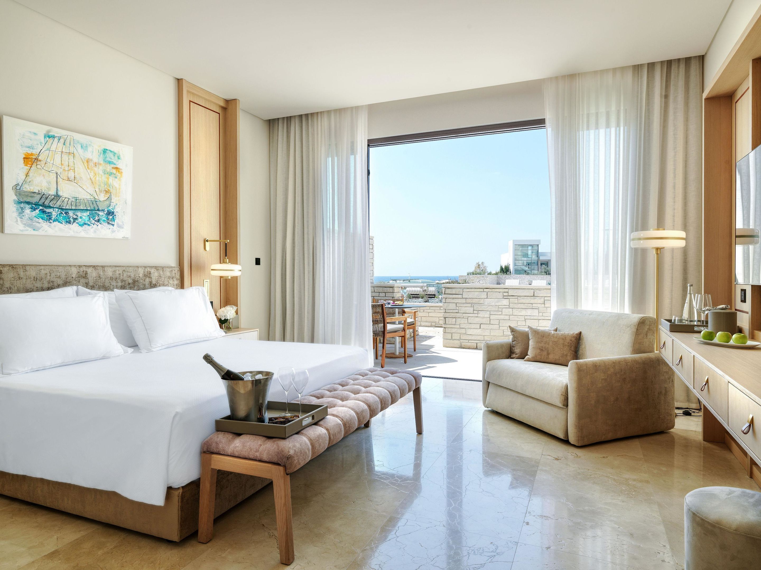A hotel room with a bed, a sitting area with a chair, a small table with a bottle and glasses, and a view of the sea.