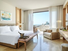 A hotel room with a bed, a sitting area with a chair, a small table with a bottle and glasses, and a view of the sea.