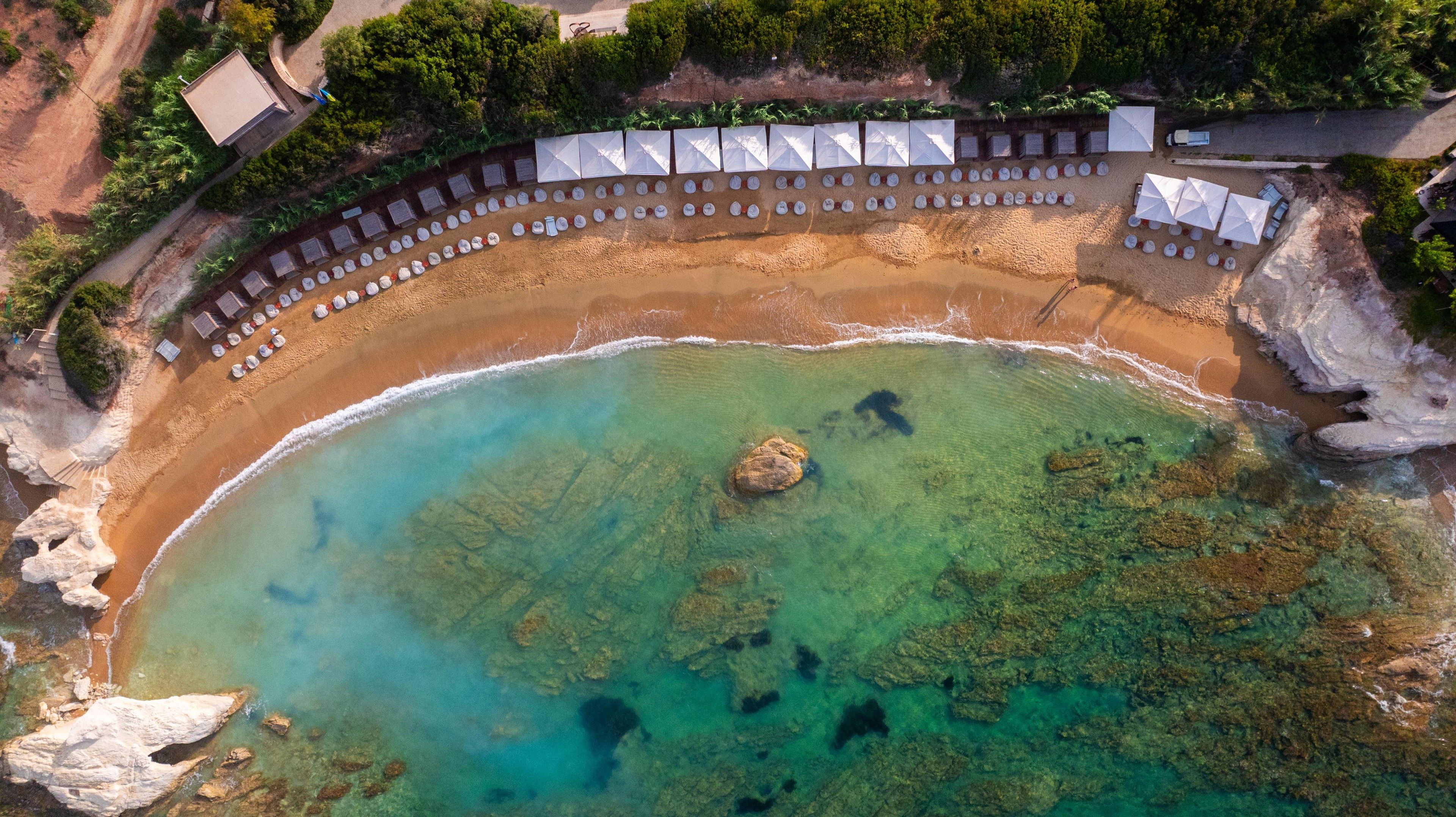 An aerial view of a beach with clear turquoise water, a rocky center, and a row of white umbrellas along the shore.