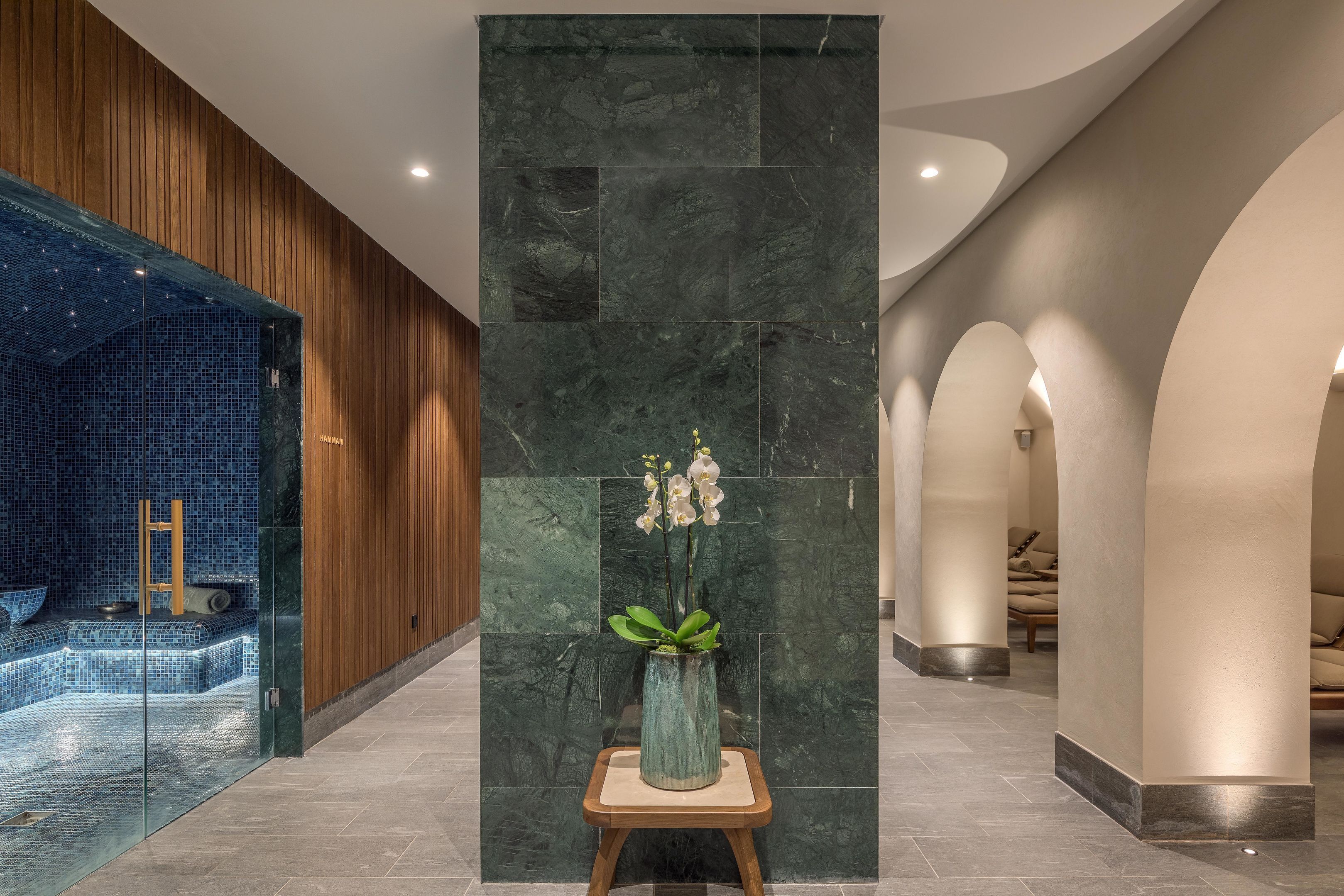 Zona de spa con pared de mosaico azul, banco de madera y un jarrón con flores blancas sobre un pedestal.
