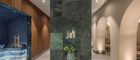 Zona de spa con pared de mosaico azul, banco de madera y un jarrón con flores blancas sobre un pedestal.