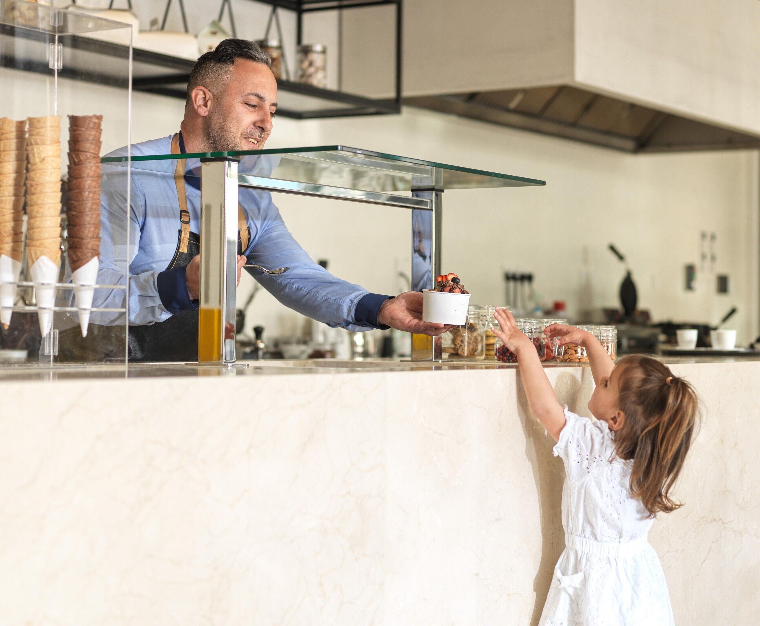 A person serving a cup of coffee to a child at a counter with various food items displayed.
