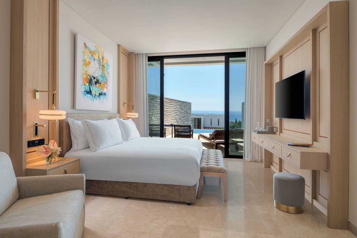 Une chambre d’hôtel moderne dotée d’un grand lit, d’un téléviseur à écran plat, d’un bureau avec une chaise et offrant une vue sur la ville grâce à de grandes fenêtres.