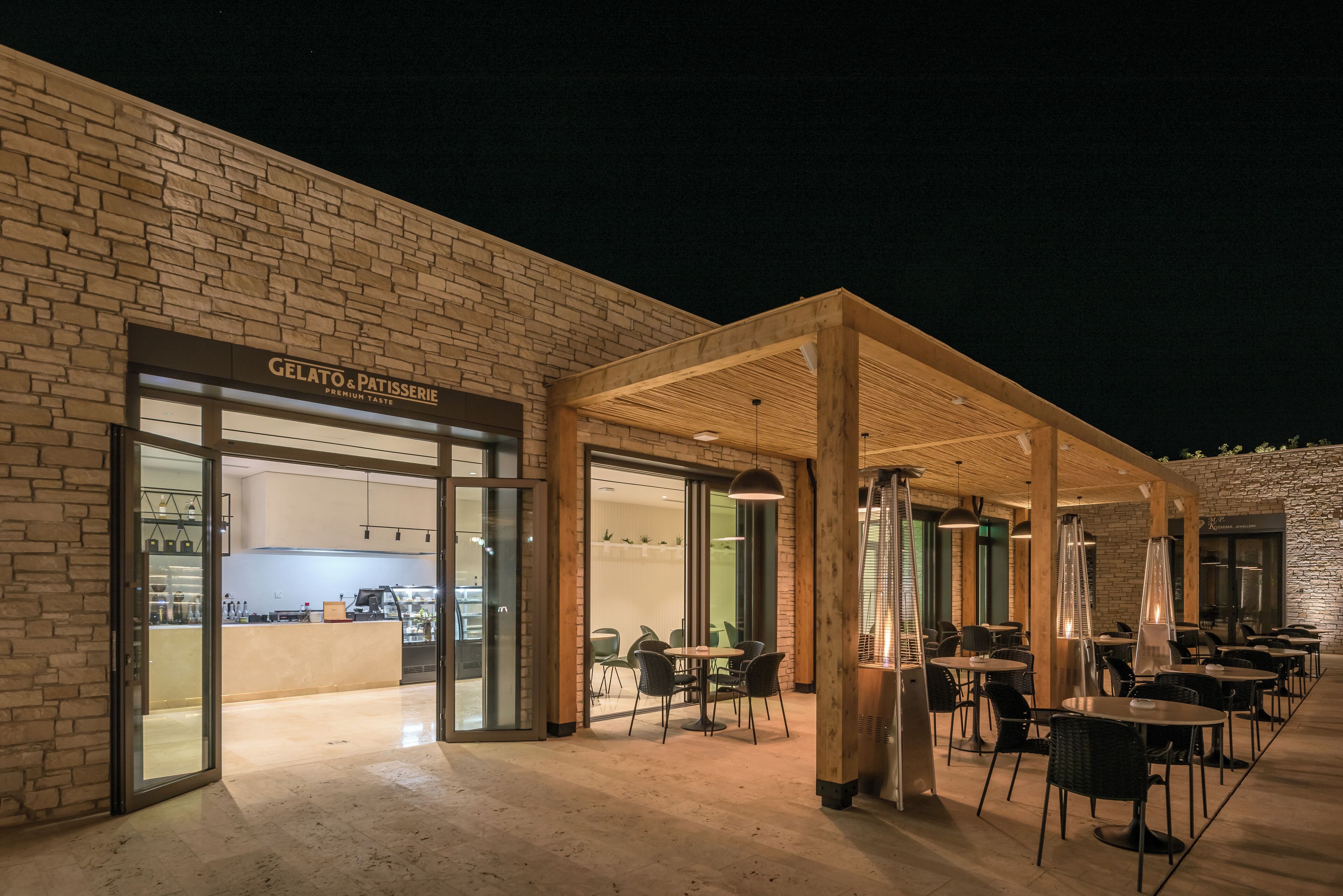Un bâtiment moderne avec une façade attrayante, un café appelé « Gelato & Patisserie » et un espace extérieur aménagé avec des tables et des chaises.