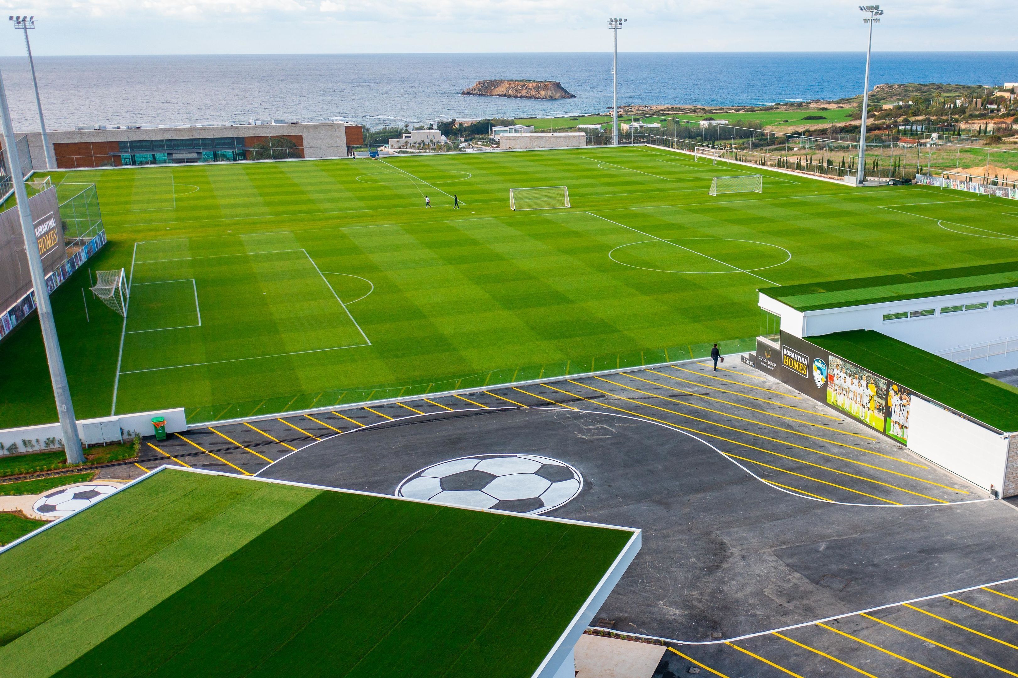 Un campo de fútbol con porterías, un estacionamiento con diseño de balón de fútbol y una vista del mar y edificios al fondo.
