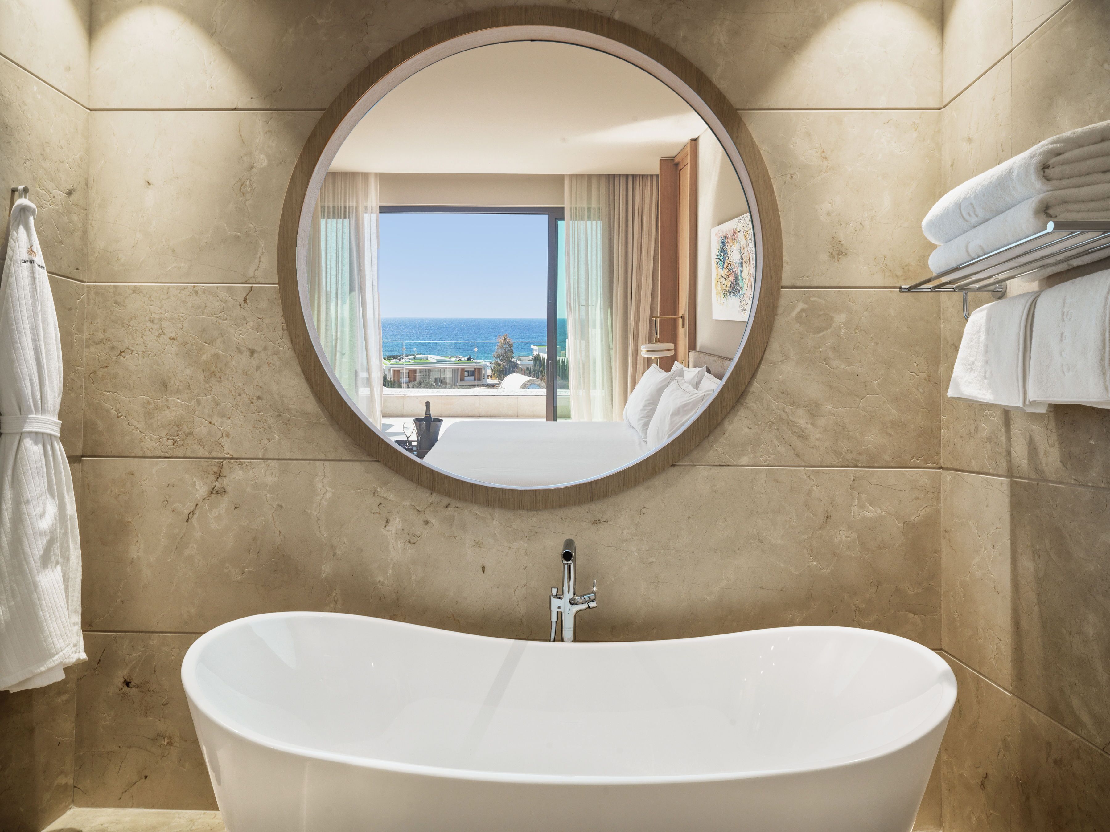 A bathroom with a freestanding bathtub, a round mirror, and a view of a balcony with a pool and ocean.