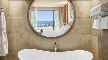 A bathroom with a freestanding bathtub, a round mirror, and a view of a balcony with a pool and ocean.