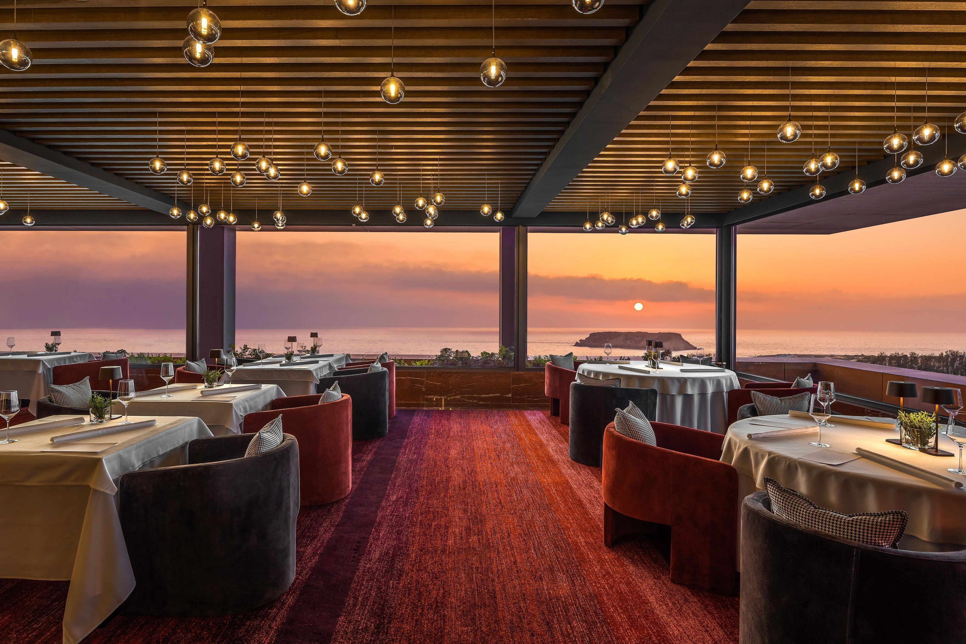 A restaurant with tables set for dining, a view of the sunset over the ocean, and hanging spherical lights.