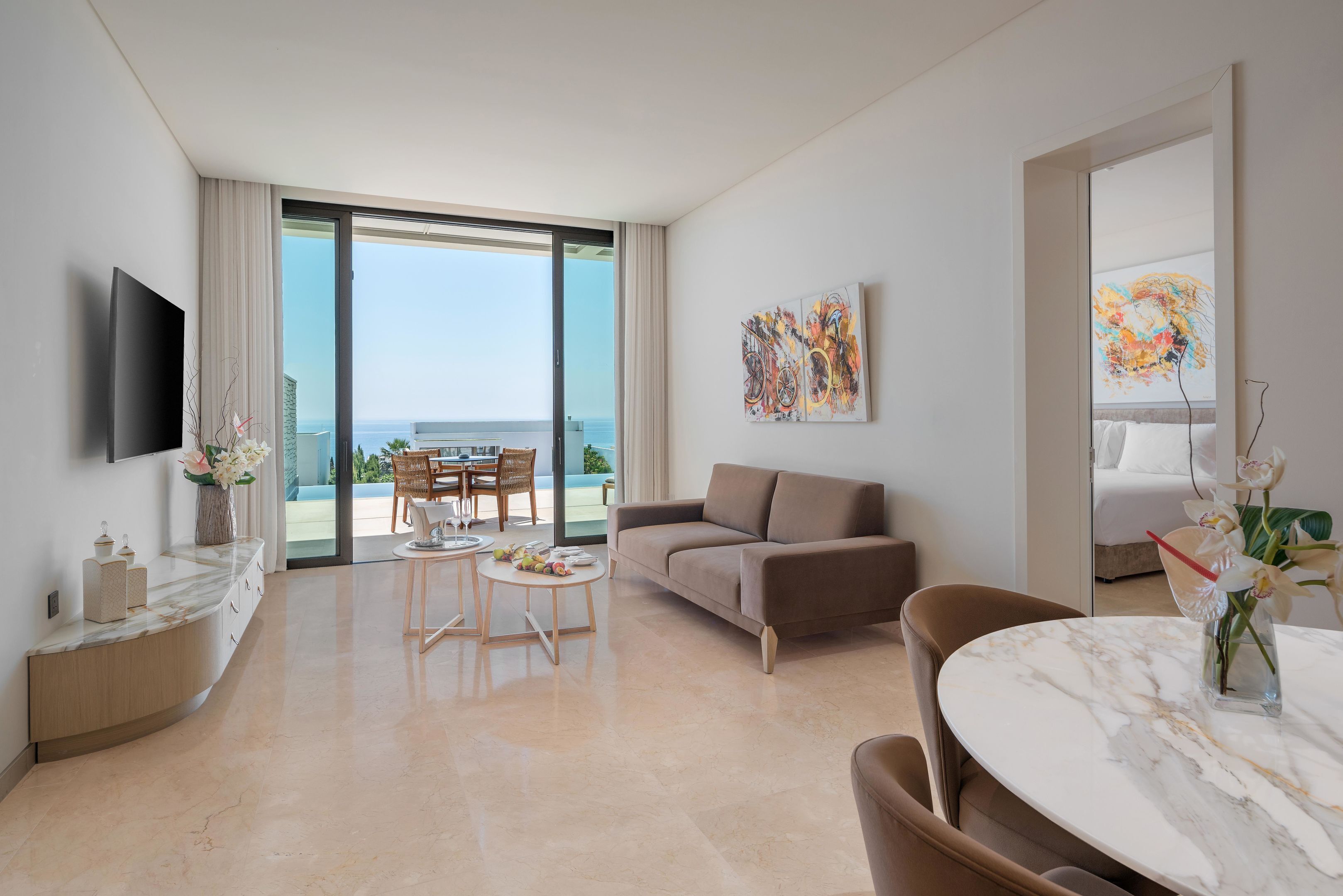 A modern hotel room with a large balcony, a flat-screen TV, a sofa, a dining area, and artwork on the walls.
