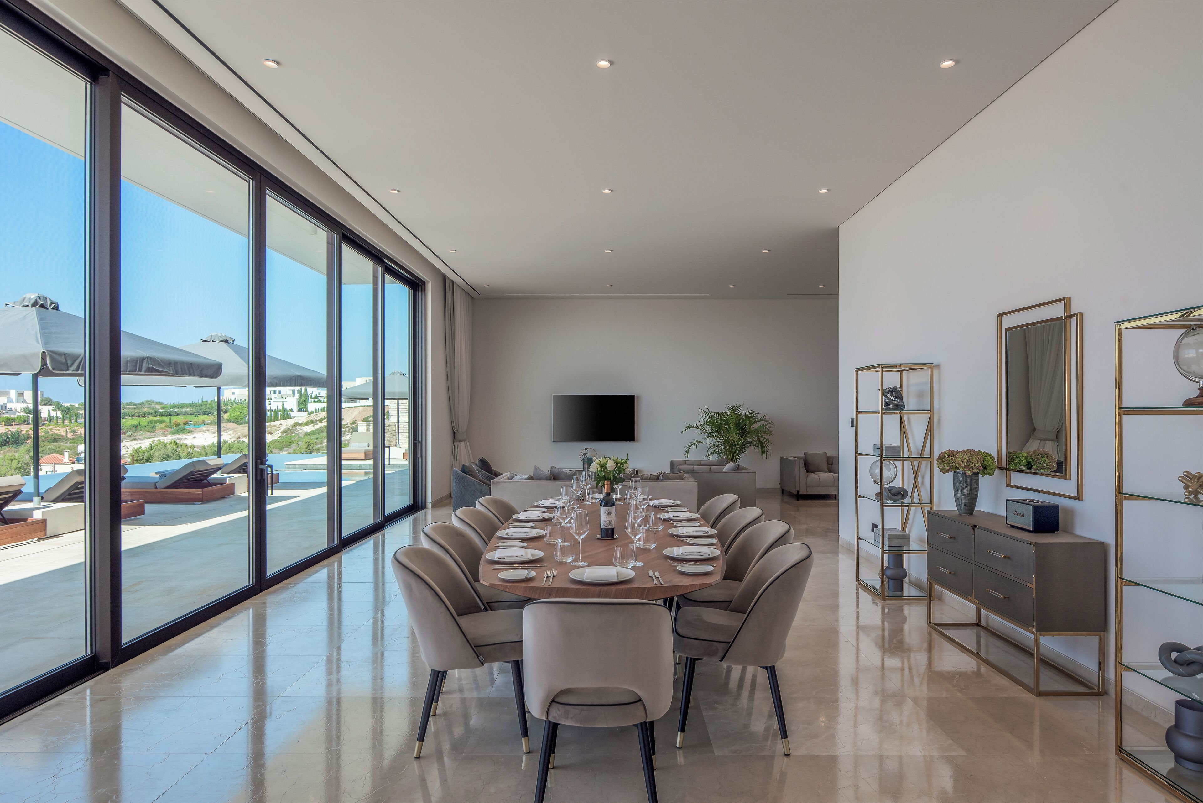 A modern dining room with a large table set for six, a flat-screen TV mounted on the wall, and a view of the outdoors through large windows.