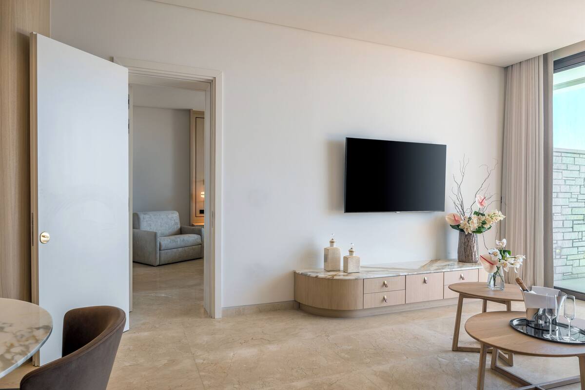 A modern living room with a flat-screen TV mounted on the wall, a round dining table with chairs, a sofa, and a cabinet with decorative items.