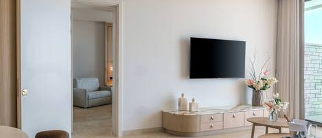 A modern living room with a flat-screen TV mounted on the wall, a round dining table with chairs, a sofa, and a cabinet with decorative items.