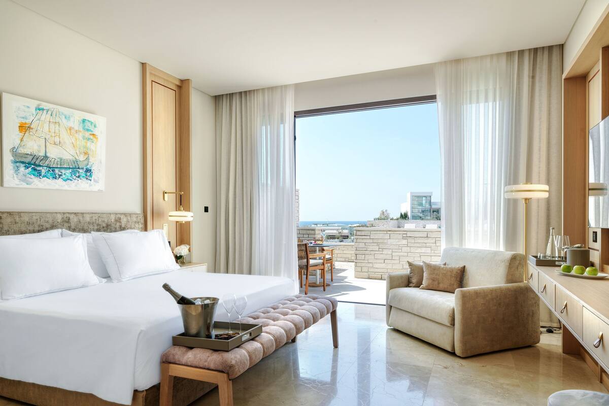 Une chambre d’hôtel comprenant un lit, un coin salon avec un fauteuil et un pouf, une table à manger avec un plateau, et une vue sur l’océan.