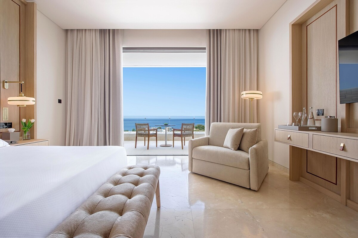 A hotel room with a large bed, a seating area with a sofa and armchair, a dining table with chairs, and a view of the sea through large windows.