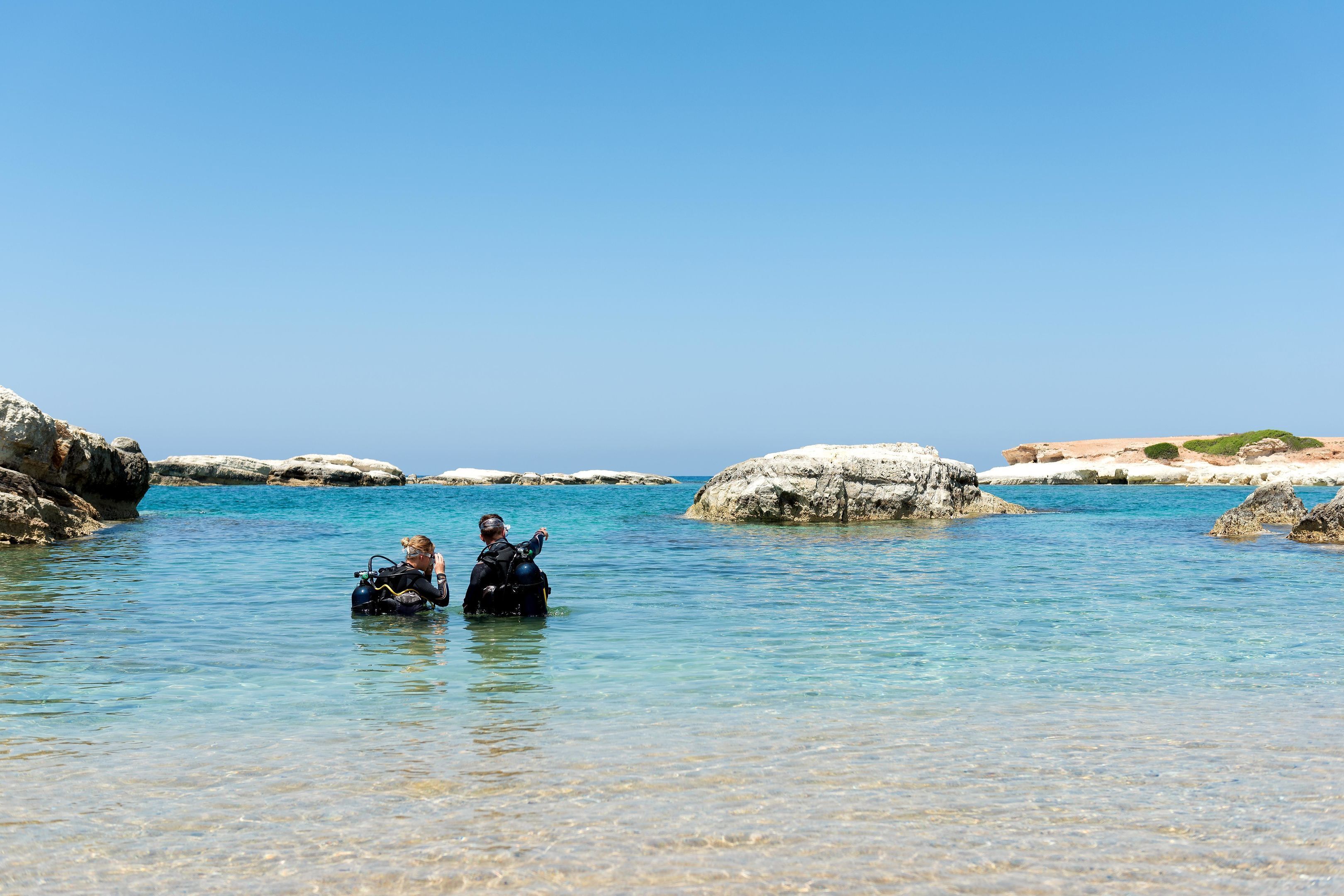 Two people in clear water near rocks, with a clear sky.