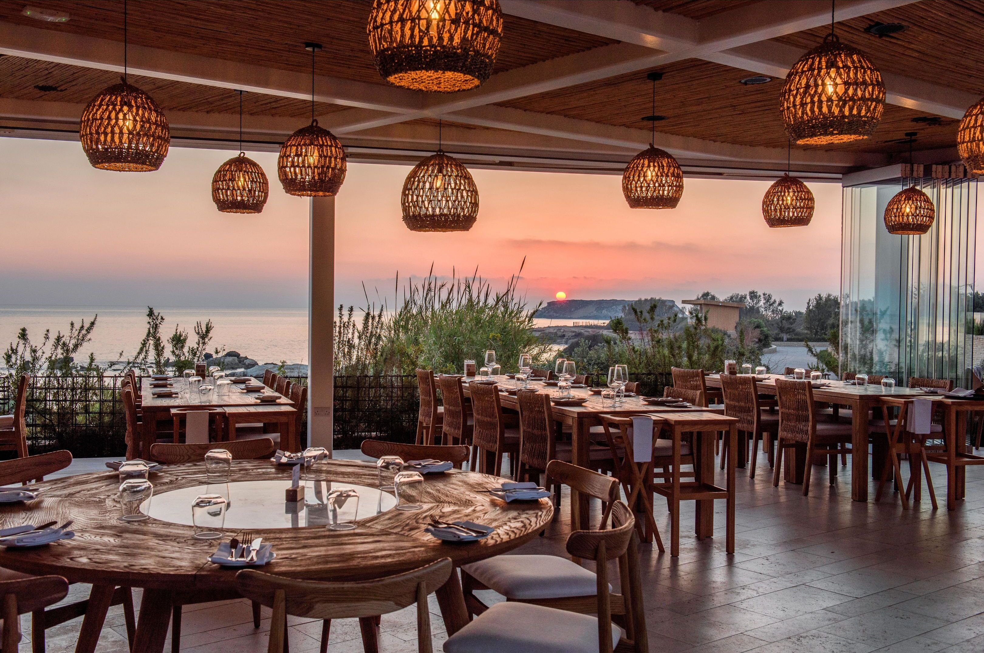 Un restaurante con mesas redondas y lámparas colgantes de mimbre, con vistas panorámicas al océano y las colinas.