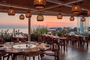 A restaurant with round tables and wicker hanging lights, overlooking a scenic view of the ocean and hills.