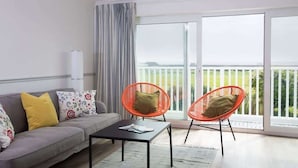 Un salon moderne avec un canapé gris, deux fauteuils orange, une table basse noire et une grande fenêtre donnant sur un paysage verdoyant.