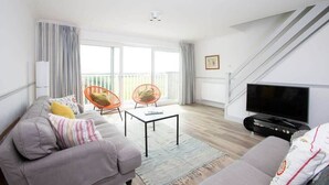 Un salon moderne avec un canapé gris, deux fauteuils orange, une table basse, un téléviseur à écran plat et un balcon avec des rideaux.