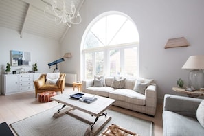 A spacious living room with a high ceiling, a large arched window, a white sofa, a leather armchair, a coffee table, and a telescope. - West Wittering Beach House for 6 (West Wittering)