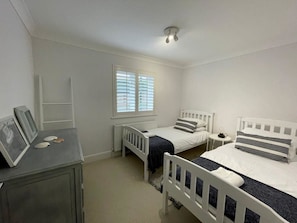 A bedroom with two single beds, a dresser, a window with blinds, and a ceiling light. - West Wittering Beach House for 6 (West Wittering)